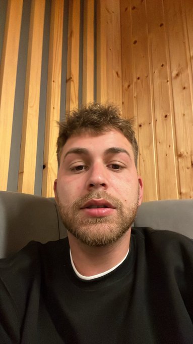 A young man with spiky brown hair and a short beard sits on a gray couch against a wood-paneled wall, appearing to address the camera in a selfie-style video inviting viewers to drop their $SOL (Solana) crypto wallet addresses for a morning giveaway.