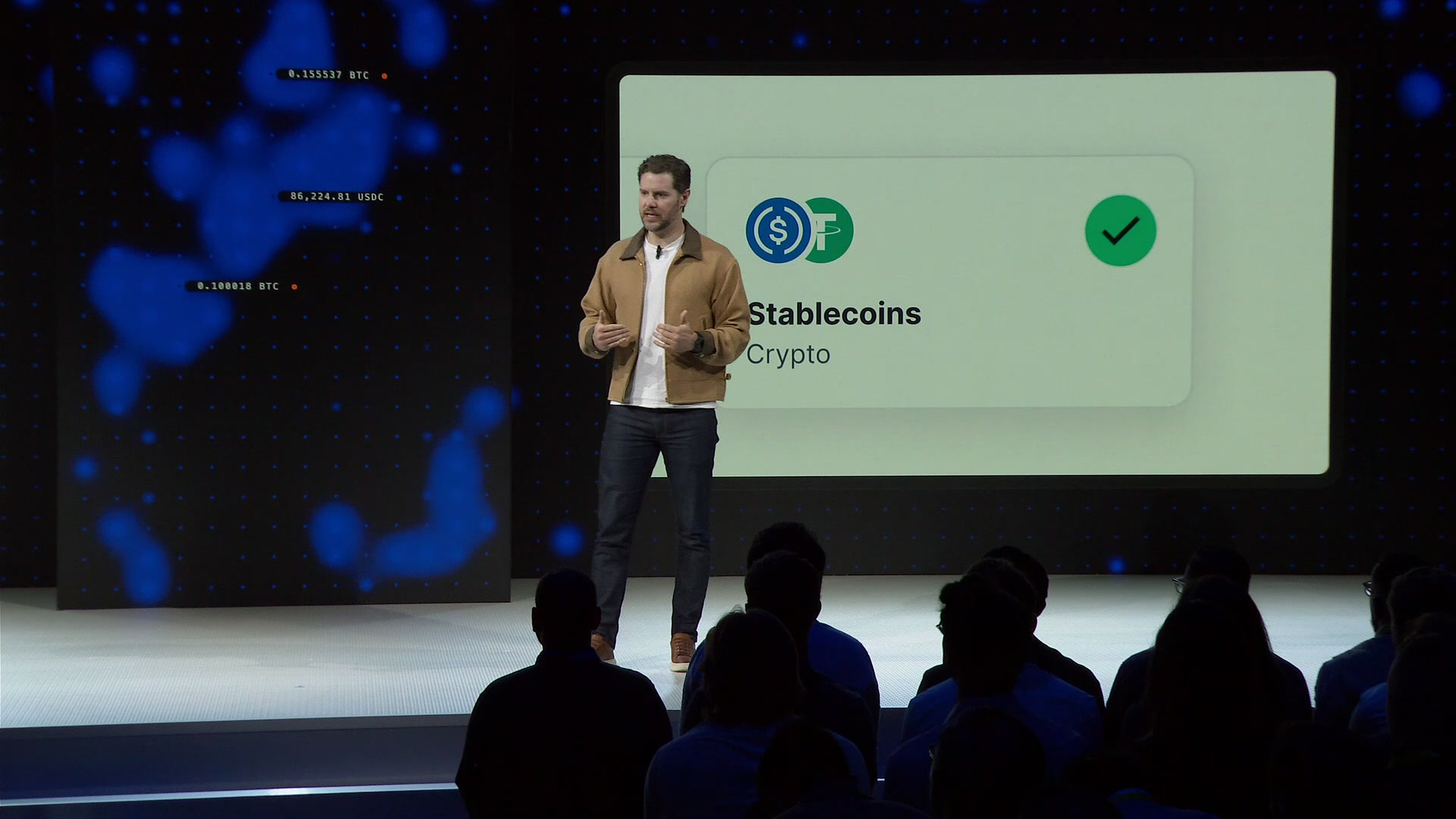 The image shows a man standing behind a podium with a screen displaying information related to cryptocurrency. The man appears to be giving a presentation or speech about the future of payments using Bitcoin as an example. There are several people sitting in front of him, attentively listening and watching his words. The room is filled with blue lights that illuminate the scene, creating a focused atmosphere for the event.