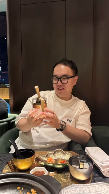 A man wearing glasses and a white "ESSENTIALS" t-shirt smiles while holding a small brass teapot at an elegant dinner table set with assorted dishes and bowls. The caption reflects his anniversary of joining Bybit, capturing a warm, celebratory moment as he marks seven years with the company.