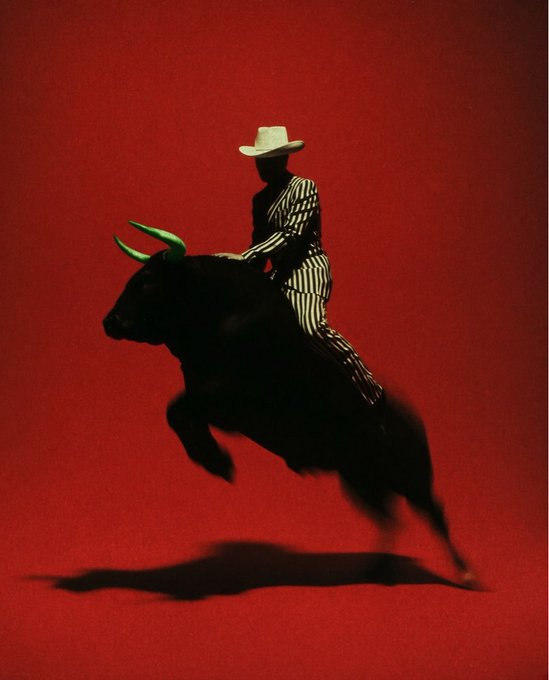 A striking artistic image of a silhouetted figure in a striped outfit and white cowboy hat riding a black bull with green-tipped horns, set against a solid, vivid red background with a shadow cast below.