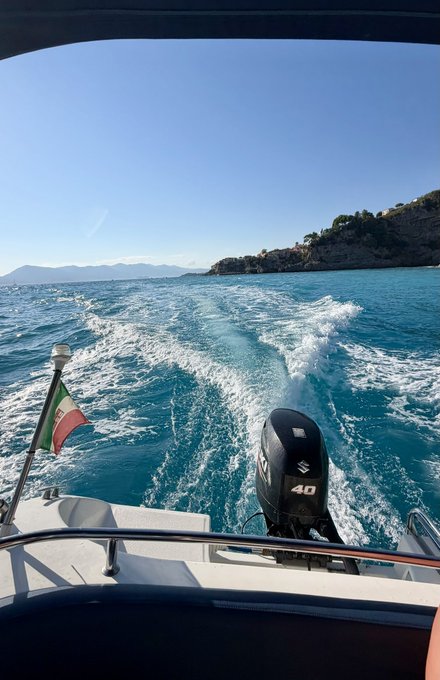 A view from the back of a boat traveling across turquoise water, showing the wake trailing behind a 40-horsepower Suzuki outboard motor, with an Italian flag flying and a rocky coastline visible in the distance under a clear blue sky.