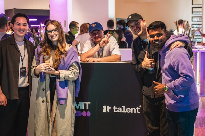 Five young people pose for a photo at an event, standing behind a black sign that reads 'talent' with part of another logo visible, suggesting a tech or company gathering.