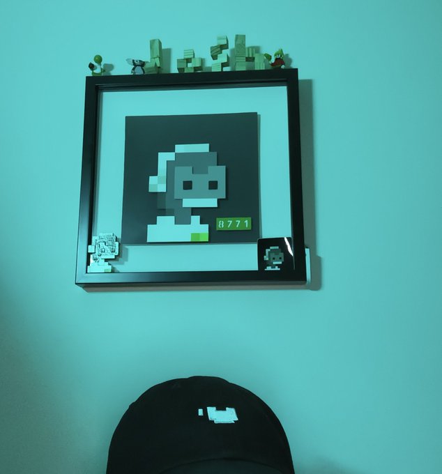 A framed pixel art portrait of a figure with the number '0771' hangs on a teal wall, topped with small blocky figurines, above a person wearing a black cap with a pixel design.