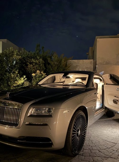 A luxury Rolls-Royce convertible parked at night with its door open, illuminated interior visible, displayed on a stone driveway with greenery and a building in the background.