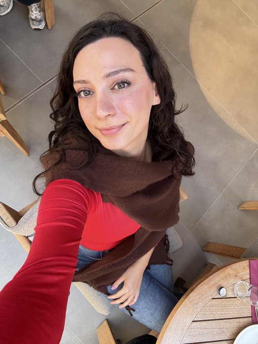 A smiling woman with brown hair sits at a table, wearing a red long-sleeve top, blue jeans, and a brown scarf draped around her shoulders.