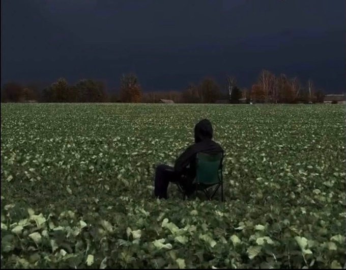 A hooded figure sits in a green folding chair in the middle of a vast green field at dusk or night.