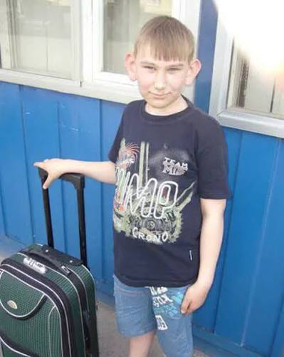 A young boy in a black graphic t-shirt and denim shorts stands next to a green rolling suitcase in front of a blue wall.