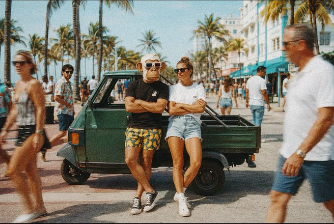 The image shows a man and woman standing next to a green truck, posing for a photo with their arms crossed. They are wearing shorts and sunglasses. In the background, there are other people walking around, some of whom appear to be carrying backpacks or handbags. The scene suggests that they might be enjoying a day at the beach or a similar outdoor location.