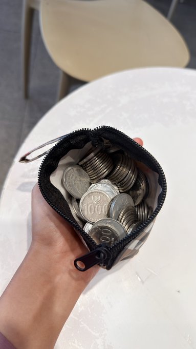 A hand holds open a small black coin purse filled with South Korean 100 won coins, with a white table in the background.