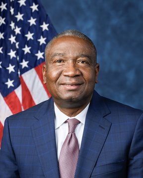A professional portrait of a smiling man in a blue suit with a pink patterned tie, photographed in front of an American flag against a blue backdrop. The formal headshot is paired with a caption suggesting irony about someone's well-timed actions or predictions.