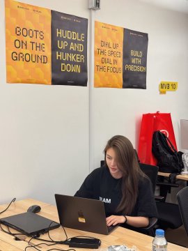A woman works on a laptop in an office space adorned with motivational banners reading "BOOTS ON THE GROUND," "HUDDLE UP AND HUNKER DOWN," "DIAL UP THE SPEED DIAL IN THE FOCUS," and "BUILD WITH PRECISION," alongside a Binance-themed "Happy 9" decoration. The setting reflects the post's nostalgic reflection on the dedication, late nights, and lessons learned behind a crypto career, embodying the perseverance behind the phrase "故事开始于一次相信" (the story began with a belief).