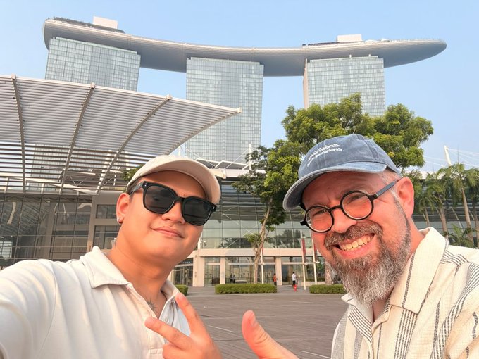 A selfie of two smiling men wearing caps and sunglasses in front of the iconic Marina Bay Sands hotel in Singapore, with one giving a peace sign and the other a thumbs-up.