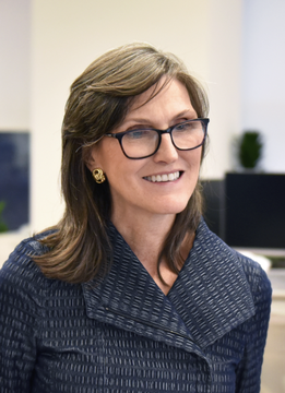 The image features an older woman wearing glasses and a blue jacket with a gold earring. She is smiling at the camera while standing against a white wall, which serves as the background for her portrait. The photo also includes text that reads "CATHIE WOOD CALLS FOR $750K BITCOIN BUT SELLS ITS OWN BTC ETF ARK Invest’s long-term $750,000 bull case still points to a massive $750,000 target." This suggests that the woman might be involved in cryptocurrency trading or investing.