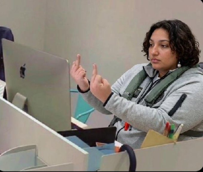 A woman in a gray hoodie sits at a desk using an Apple MacBook laptop, both middle fingers raised in a defiant gesture. She appears to be in an office setting, matching the meme's tone of someone defiantly dismissing skepticism about a modest investment turning into significant gains.