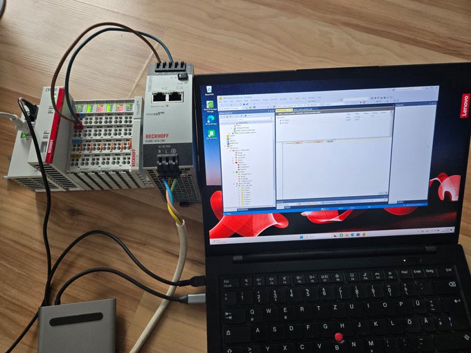 A Lenovo laptop displaying what appears to be TwinCAT PLC programming software is connected via Ethernet cable to a Beckhoff PLC system with terminal blocks, set up on a wooden desk.