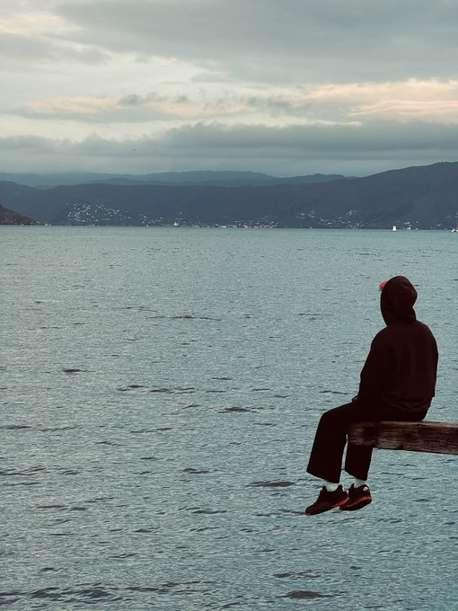 A serene photograph of a hooded figure wearing a red beanie sitting on a wooden plank extending over a calm body of water, gazing toward distant mountains under an overcast, cloudy sky.