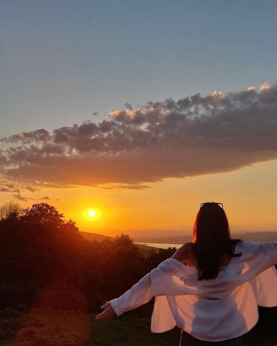 A woman in a white top stands with arms outstretched, watching the sunset over a landscape of trees and water.