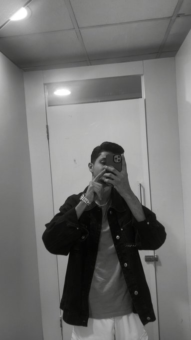 A black-and-white mirror selfie of a man wearing a dark denim jacket and white pants, holding a phone to cover his face while wearing a watch and bracelet.