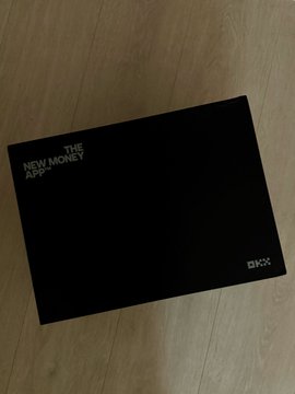 A black album cover or booklet on a wooden floor with text reading 'THE NEW MONEY APK' and 'OKX'.