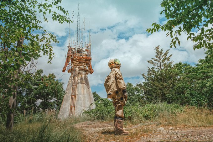 A person in a vintage-style space suit with a gold helmet stands on a rocky outcrop looking at an old, weathered rocket tower surrounded by greenery under a cloudy sky.