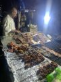 Two people are cooking skewered meat on a large charcoal grill in a dimly lit, nighttime setting.