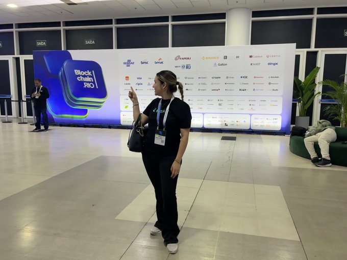 A woman wearing a lanyard poses in front of the "Block|chain|RIO" backdrop featuring sponsor logos like Coinbase, Binance, and OKX, connecting at the conference to network with builders and founders in the LATAM region seeking stablecoin infrastructure solutions.