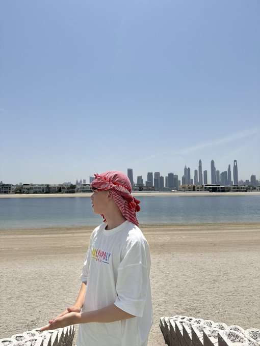 A person wearing a red keffiyeh and a white t-shirt with colorful text stands on a sandy beach with a city skyline visible across the water.