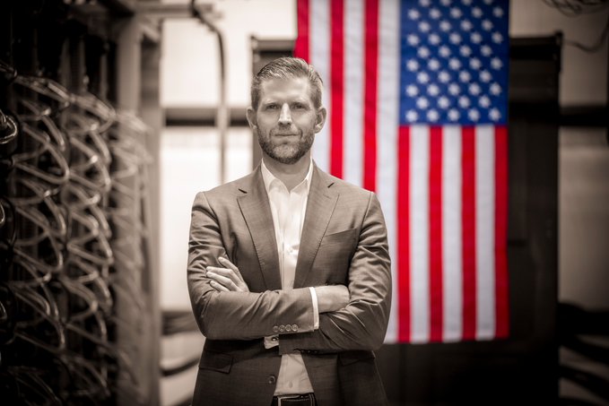 Eric Trump standing in a suit with arms crossed in front of an American flag, in a black-and-white photo with the flag in color.