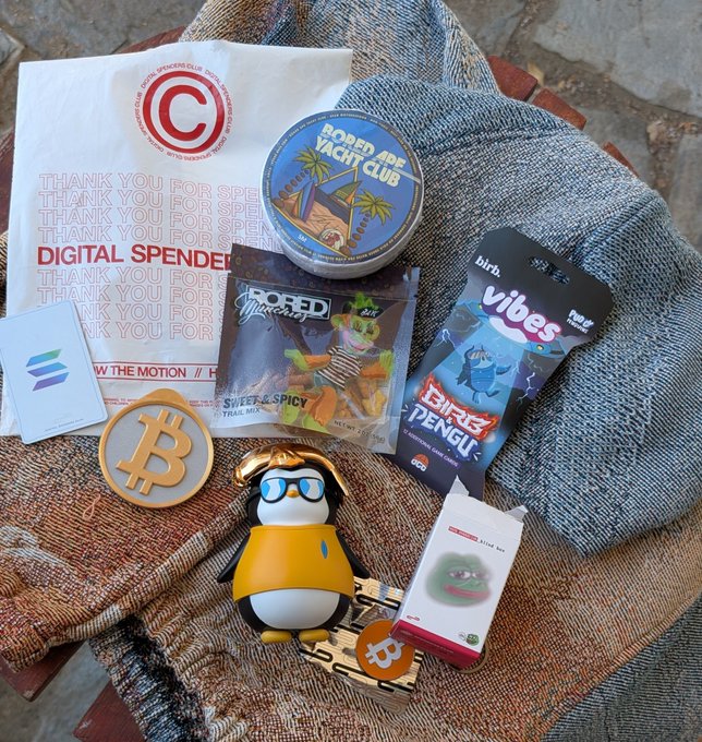 A flat-lay photo of crypto-themed merchandise including a 'Digital Spender' bag, a Bitcoin coin, a penguin figurine with sunglasses, a 'Roped Yacht Clubs' tin, Bobo'd spicy trail mix, Dingo Pengu candy, and a pink item labeled 'blink box'.