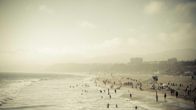 A wide, hazy view of a crowded beach with people swimming in the ocean and walking along the shoreline, with buildings visible in the misty distance.