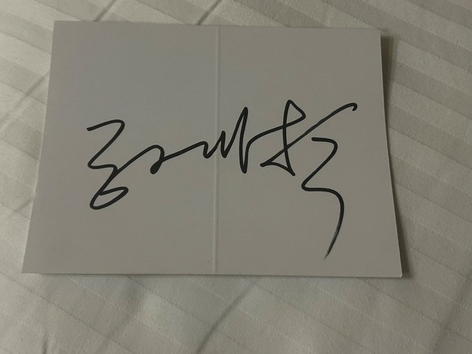 A black ink signature is written on a white folded piece of paper resting on a striped fabric surface.
