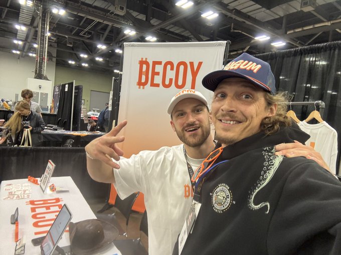 Two men take a selfie at what appears to be a trade show or convention, posing in front of a white banner that reads 'DECOY' in orange lettering.