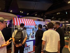 A photo of people gathered at an indoor event or convention, with a red and white striped booth visible in the background and attendees wearing casual clothing and face masks.