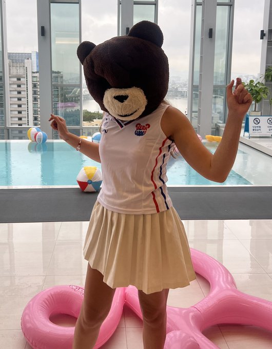 A person wearing a large brown teddy bear head, a white sleeveless top, and a beige pleated skirt poses playfully by an indoor pool with a pink flamingo float.