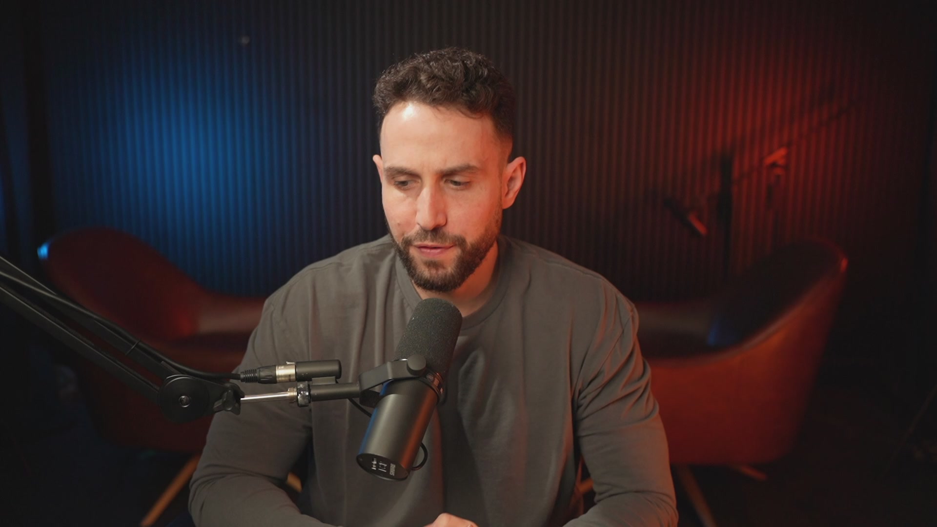 The image shows a man sitting at a microphone, possibly recording or broadcasting something. The background features a dark room with red and blue lights, creating an atmosphere of mystery or intrigue. There are two chairs visible in the scene - one on the left side and another on the right side. Additionally, there is a computer monitor placed behind the man, suggesting that he might be working on some content creation or broadcasting project. The image also contains text that reads "AI Stocks are printing 10X gains Meanwhile BTC is flashing warning signs that more downside could be coming LIVE with @gregosuri". This implies that the man may be discussing a topic related to cryptocurrency and its impact on the stock market, possibly drawing attention to potential risks or opportunities in this area.