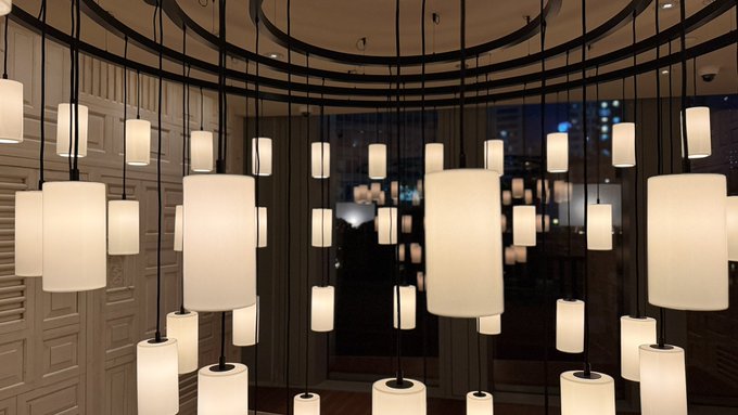 An interior view of a modern hallway or lounge featuring numerous cylindrical pendant lights hanging at varying heights against paneled walls and a large window.