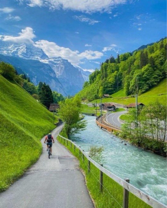 A scenic landscape shows a person biking along a paved path beside a rushing turquoise river, with green hillsides and snow-capped mountains in the background.