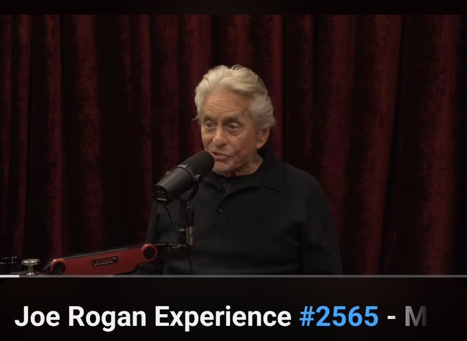 A screenshot from the "Joe Rogan Experience #2565" podcast shows an older man with white hair and a mustache speaking into a Shure microphone against a dark red curtain backdrop.