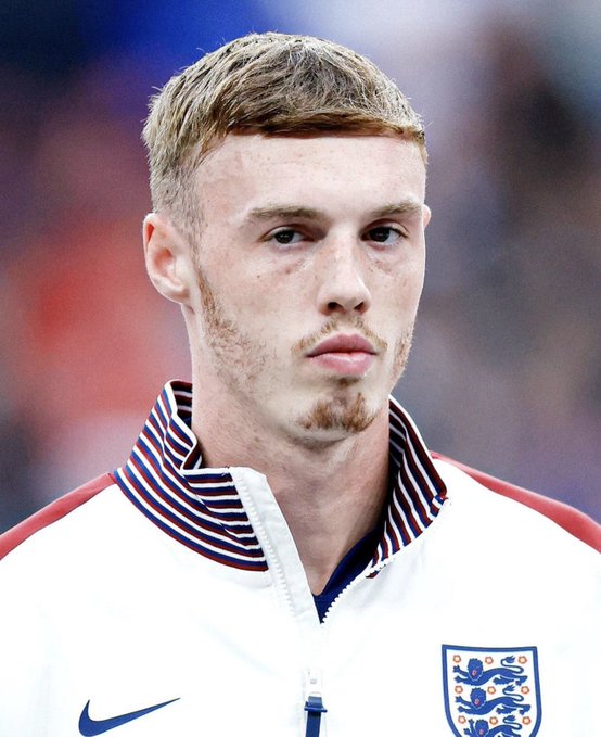 Portrait of a young man with blond hair and a small beard wearing an England national football team jacket with the Three Lions crest and Nike logo.