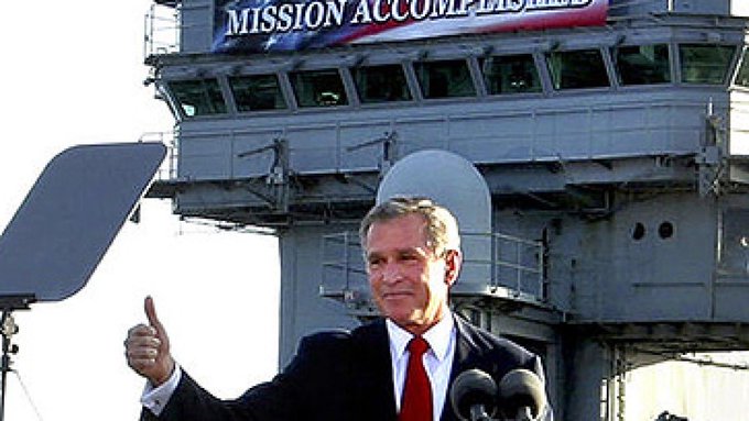 President George W. Bush gives a thumbs-up while standing in front of an aircraft carrier control tower with a 'MISSION ACCOMPLISHED' banner hanging above.