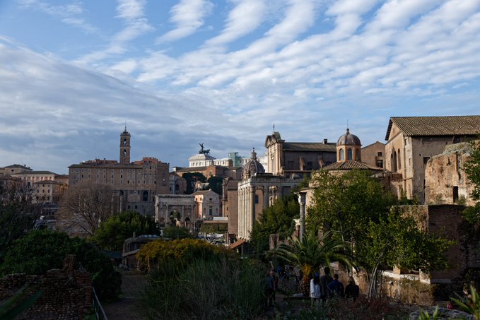 A scenic view of Rome featuring historic buildings, domes, and ancient ruins under a sky filled with wispy clouds, with greenery in the foreground.
