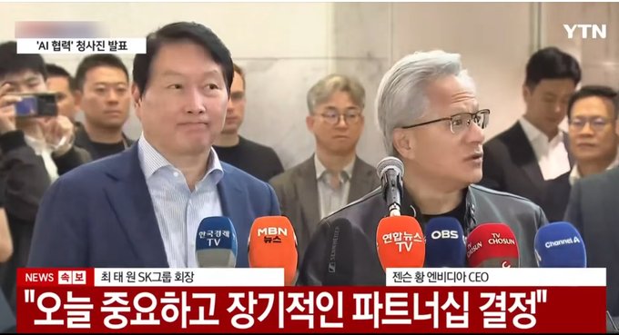 The image shows two men standing at microphones during an interview or press conference. They are both wearing suits and appear to be well-dressed for the occasion. The background of the image features a crowd of people, indicating that this event might be taking place in front of a large audience.