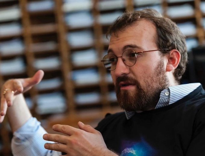 The image shows a man wearing glasses, with his hands up as if he's explaining something or making a point. The background features a bookshelf filled with numerous books, suggesting that this might be an office or a library setting where people gather for discussions or presentations. The caption reads "No protocol hacks. No major outages. No crazy fees. Constant development. Cardano keeps building while others chase hype." This implies that the man is talking about the importance of focusing on practical solutions and not getting caught up in speculative or hype-driven projects, which can lead to wasted time and resources.