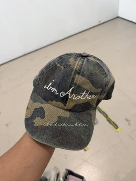 A camouflage baseball cap with the embroidered text 'I'm Another' is held up against a tiled floor background.