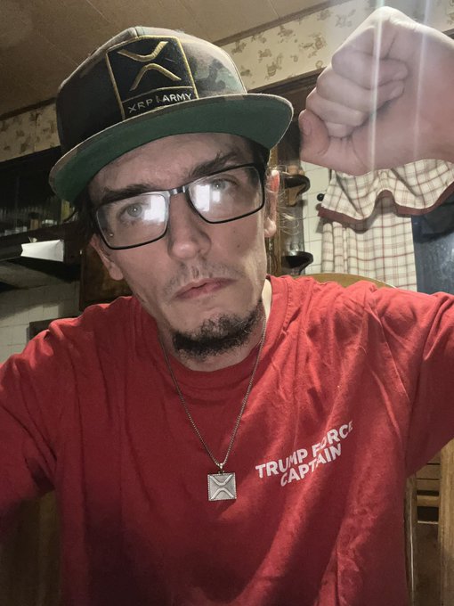 A selfie of a man wearing an XPR Army cap, glasses, a Trump Force Captain red t-shirt, and an XPR pendant necklace.