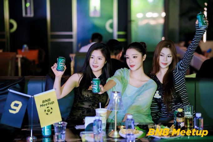 Three women at a 'BNB Meetup' event hold up green BNB-themed cans at a table marked with a '9' and a small 'BNB Meetup' flag.