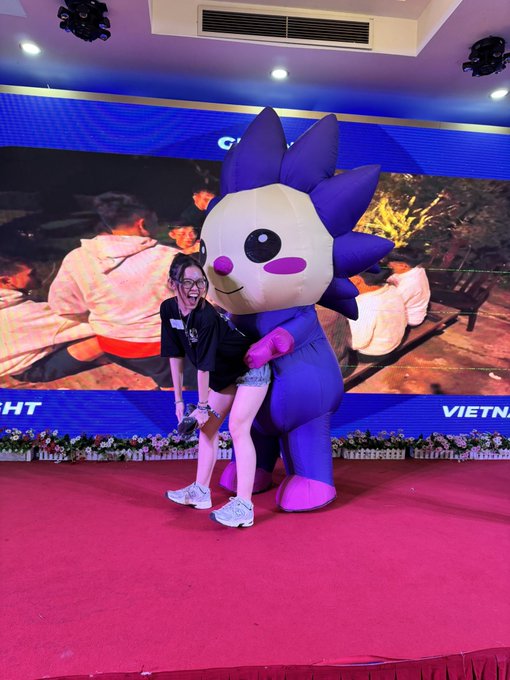 A young woman poses playfully with a purple mascot character on a stage featuring a red carpet and a backdrop screen displaying "VIETN..." text, promoting the upcoming NFT mint date of 28-08-2026. The event setting, with its mascot and branded backdrop, suggests an exciting launch gathering to engage the community ahead of the big reveal.