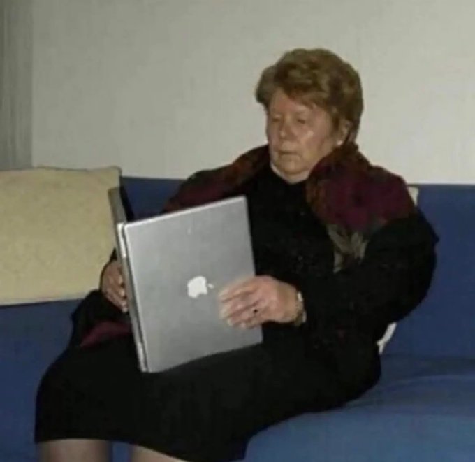 A grainy photo of an older woman sitting on a blue couch, awkwardly holding a silver Apple laptop while looking confused.