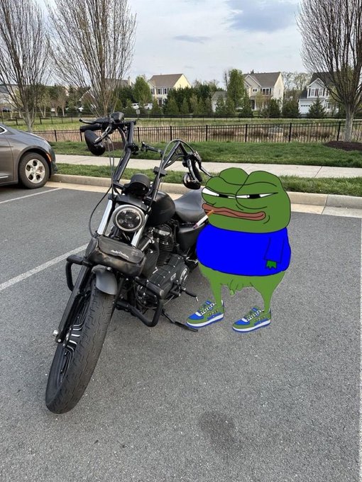 A humorous edited photo showing the Pepe the Frog meme character wearing a blue shirt and sneakers standing next to a black motorcycle in a parking lot.