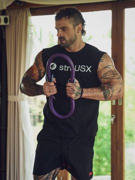 A tattooed man in a sleeveless "strcUSX" shirt demonstrates a stretch using a purple resistance ring, visually tying into the post about learning about strcUSX at Solstice's stretching event. The casual indoor setting with tropical greenery through the window complements the relaxed, instructional tone of combining fitness with DeFi education.